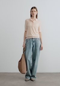 Ανοιχτό ροδακινί κοντομάνικο πόλο πουκάμισο με κουμπιά, συνδυασμένο με φαρδιά ανοιχτά μπλε τζιν. Το μοντέλο κρατά μία καφέ τσάντα tote και φοράει μπεζ παπούτσια.