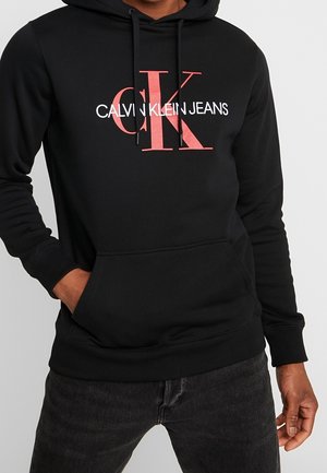 Person trägt schwarzen Hoodie mit rotem "CK"-Logo und weißem "CALVIN KLEIN JEANS" Schriftzug auf der Brust, Hände in den Vordertaschen.