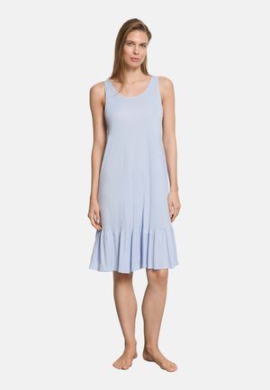 Femme debout pieds nus portant une robe bleu clair sans manches longueur genou avec un ourlet volanté sur un fond blanc uni.