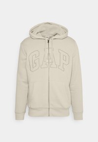 Sweat à capuche beige zippé avec intérieur pelucheux, arborant un grand logo "GAP" en relief sur le devant. Comprend deux poches avant et des poignets côtelés.