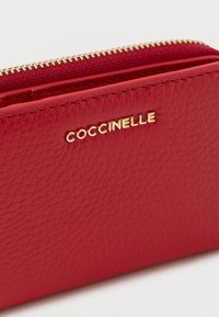 Punainen nahkalompakko teksturoidulla pinnalla, jossa on kullanvärinen vetoketju ja vastaava kohokuvioitu logo. Kompakti muotoilu, jossa korttipaikat.