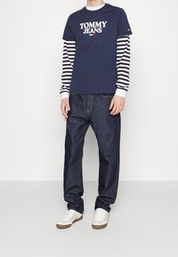 Námořnické tričko s krátkým rukávem značky Tommy Jeans přes bílé a černě pruhované tričko s dlouhým rukávem, v kombinaci s tmavě modrými džínovými kalhotami a bílými teniskami.