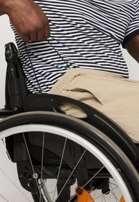 Personne portant une chemise rayée et un pantalon beige assise dans un fauteuil roulant, tenant la roue pour se propulser vers l'avant.