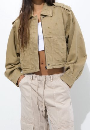 Vrouw draagt een cropped, versleten beige jas, wit cropped topje en lichtbeige broek met koord, handen in de zakken.