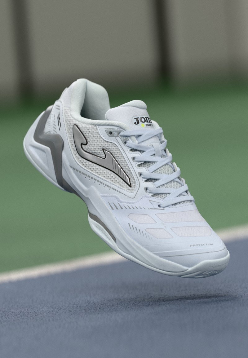 Hvide og grå Joma sportsko svævende over en blå tennisbane med en grøn baggrund.