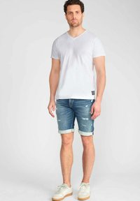 Witte katoenen V-hals t-shirt, blauwe denim shorts met opgerolde mouwen, en witte sneakers met een lichtbruine zool. Beschadigde details op de shorts.