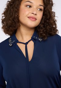 Blusa blu navy con scollatura a goccia, colletto decorato con cristalli chiari e texture liscia. Maniche lunghe e vestibilità rilassata.