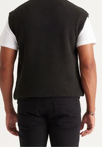 Maglione senza maniche di colore verde scuro lavorato a maglia sopra una maglietta bianca, abbinato a jeans neri. Testura a coste visibile e vestibilità casual.
