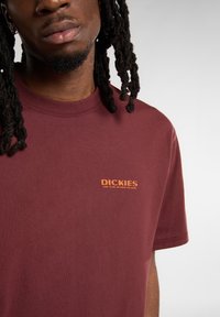 Bordová bavlněná tričko s kulatým výstřihem a krátkými rukávy, s oranžovým vyšitým logem na levé části hrudi.