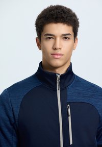 Giacca zip-up navy con colletto alto, caratterizzata da pannelli testurizzati di un blu più chiaro. Dettagli con zip e design aderente. Tessuto morbido e elastico.