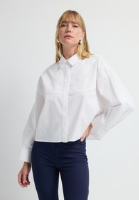 Camisa blanca de grandes dimensiones con cuello puntiagudo, cierre de botones y detalle de costura en el pecho, combinada con pantalones ajustados azul marino. Textura de tela suave.