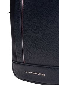 Tummansininen nahkareppu, jossa on teksturoitu pintakäsittely, pystysuorat punaiset ja valkoiset raidat sekä hopeinen logo pohjassa.