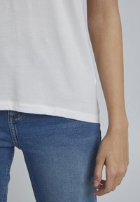 T-shirt bianco in cotone con scollatura rotonda, abbinato a jeans in denim blu. La T-shirt ha una consistenza liscia e una vestibilità leggermente ampia.