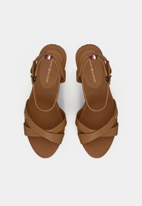 Sandales à brides croisées marron avec une semelle texturée, dotées d'une boucle en métal et d'un logo doré. Le design est élégant et minimaliste.