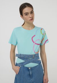 T-shirt turchese con linee astratte colorate; maniche corte e scollatura rotonda. Abbinato a una gonna di denim, caratterizzata da un design con cintura.