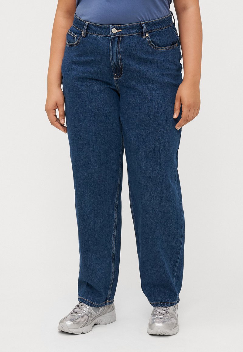 Une personne se tient debout, portant une chemise bleue et un jean bleu ample, associés à des chaussures de sport argentées, devant un fond neutre.