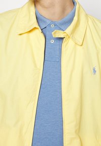 Gelbe Zip-Jacke mit Kragen über einem blauen Poloshirt. Die Jacke hat eine glatte Textur und verfügt über ein kleines Logo auf der Brust.