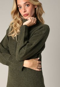 Femme aux longs cheveux blonds portant un pull en tricot vert foncé, reposant sa main sur son menton et tenant son autre bras contre son corps.