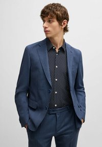 Granatowy garnitur o teksturowanym wykończeniu, z jednorzędową marynarką, klapami w szpic i bocznymi kieszeniami. Pod spodem czarna koszula w białe kropki.