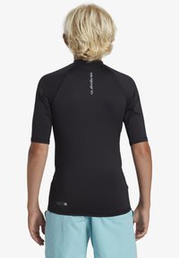 Rash guard noir à manches courtes avec une texture lisse et des coutures à plat, arborant un petit logo dans le dos, associé à un short bleu clair.