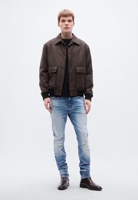 Καφέ σακάκι από μείγμα μαλλιού με κλείσιμο με κουμπιά και τσέπες-μπαλώματα, σε συνδυασμό με ανοιχτό μπλε τζιν με φθορές και σκούρα δερμάτινα μποτάκια.