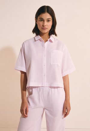 Giovane donna che indossa una camicia rosa chiaro a maniche corte con texture e bottoni, e pantaloni larghi abbinati, in piedi davanti a uno sfondo beige uniforme.