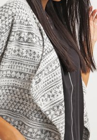 Stickad omfångsjacka i svart och vitt, med intrikata geometriska mönster, öppen framsida och mjukt, texturerat tyg.