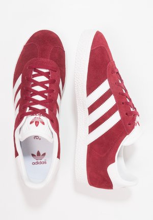 Een paar rode Adidas suède sneakers met witte strepen en zolen, gezien van boven en van opzij op een witte achtergrond.