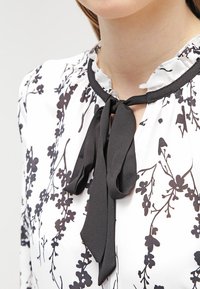 Vit blus med svart blommönster, med en volangkrage och ett svart band vid kragen. Slät tygstruktur.
