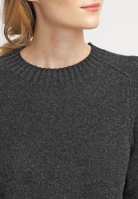 Ciemnoszary sweter wykonany z teksturowanej dzianiny, z prążkowanym okrągłym dekoltem i rękawami raglanowymi. Zbliżenie na obszar torsu.