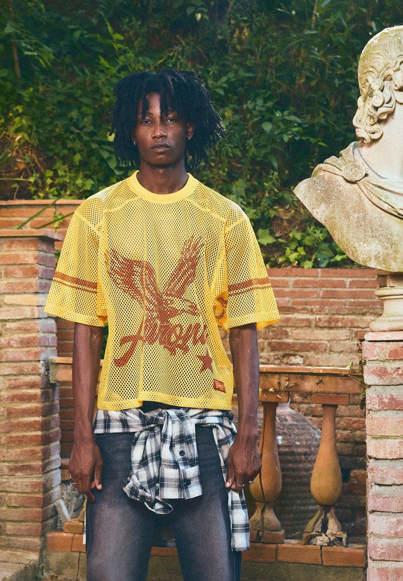 Jeune homme avec des dreadlocks portant un t-shirt en maille jaune avec un motif d'aigle et une chemise à carreaux nouée autour de sa taille, debout près d'un buste en pierre et d'un mur en briques.