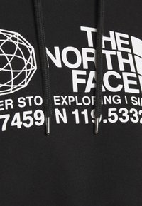 Tissu noir avec texte imprimé blanc "THE NORTH FACE", logo géométrique, coordonnées partielles et cordons de capuche noirs avec embouts métalliques.