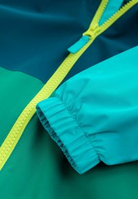 Secțiune de jachetă colorată cu țesătură turcoaz și albastru închis, cu un fermoar galben strălucitor și manșete elastice la mânecă.