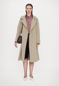 Beżowy trencz z dużym kołnierzem i srebrnymi elementami, noszony na wzorzystej brązowej koszuli, zestawiony z czarnymi szortami i kremowymi wsuwanymi butami.