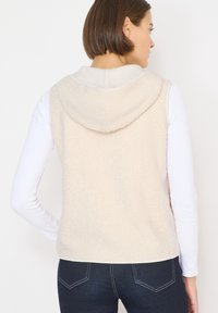 Fleece-Weste in hellem Beige mit weicher Textur, mit Kapuze und lockerem Sitz. Getragen über einem weißen Langarmshirt und dunklen Jeans.