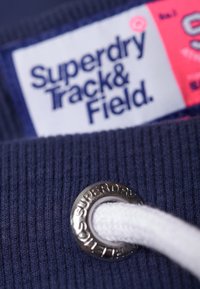 Nahaufnahme eines marineblauen Stoffs mit weißem Kordelzug durch eine Metallöse, auf der "Superdry" eingraviert ist, und einem teilweise sichtbaren Etikett mit der Aufschrift "Superdry Track & Field".