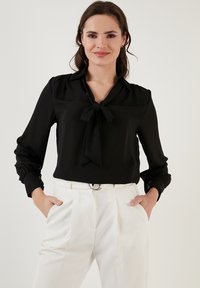 Black blouse with a tied neck, long sleeves with gathered cuffs, and a smooth texture. Worn with white tailored pants featuring a belt.