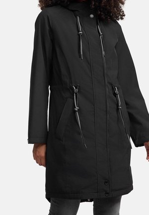 Femme portant un long manteau noir à capuche avec poches avant et cordons de serrage à la taille, debout, les bras détendus le long du corps.