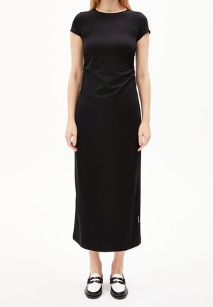 Femme portant une robe mi-longue noire unie à manches courtes et des mocassins noirs et blancs, debout devant un fond blanc.