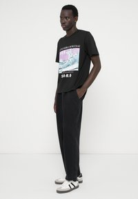 Svart grafisk t-shirt med Hokusai-vågmönster i blått och lila, parat med svarta byxor och vita skor med grå detaljer.