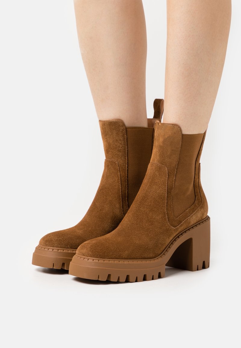 Bottes chelsea en daim marron avec talons carrés épais et pans latéraux élastiques portées sur jambes nues.
