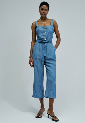 Femme debout portant une combinaison en jean bleu sans manches avec des boutons à l'avant et une ceinture à la taille, associée à des sandales à talons noires.