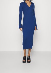 Abito lungo e aderente realizzato in tessuto a maglia a coste, con scollatura a V profonda, maniche lunghe con polsini a campana, in un vivace colore blu.