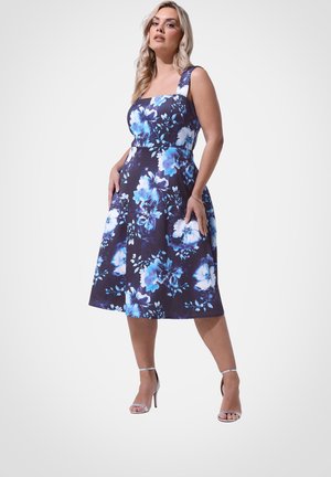 Kvinde iført en ærmeløs marineblå kjole med blåt og hvidt blomsterprint, stående med hænderne i lommerne, iført sølvfarvede høje hæle sandaler.