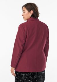 Kvinde med kort brunt hår iført en bordeaux blazer og nederdel med sorte pailletter, stående sidelæns mod en ensfarvet hvid baggrund.