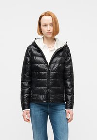 Νεαρό άτομο με ίσια κόκκινα μαλλιά, που φοράει μαύρο μπουφάν, λευκό πουκάμισο με γιακά, κολιέ με μαργαριτάρια και μπλε τζιν, σε απλό φόντο.