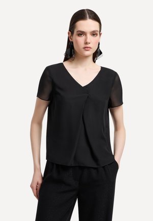 Blusa nera con maniche corte trasparenti, scollatura a V e dettaglio con piega frontale. Tessuto morbido con una texture liscia. Indossata con pantaloni neri.