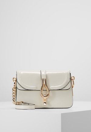 Petit sac à main couleur crème avec une fermeture dorée et une bandoulière chaîne, posé sur une surface blanche sur un fond uni.