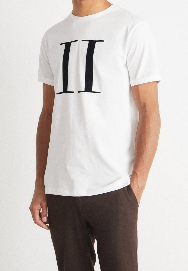 Camiseta blanca de algodón con un destacado estampado negro de "II" en la parte frontal. Cuello redondo, mangas cortas y corte relajado. Combinada con pantalones marrones.