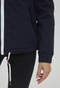 Marineblå jakke med hvit glidelås og snor, med en sidelomme og elastiske mansjetter, laget av et glatt, lett stoff.